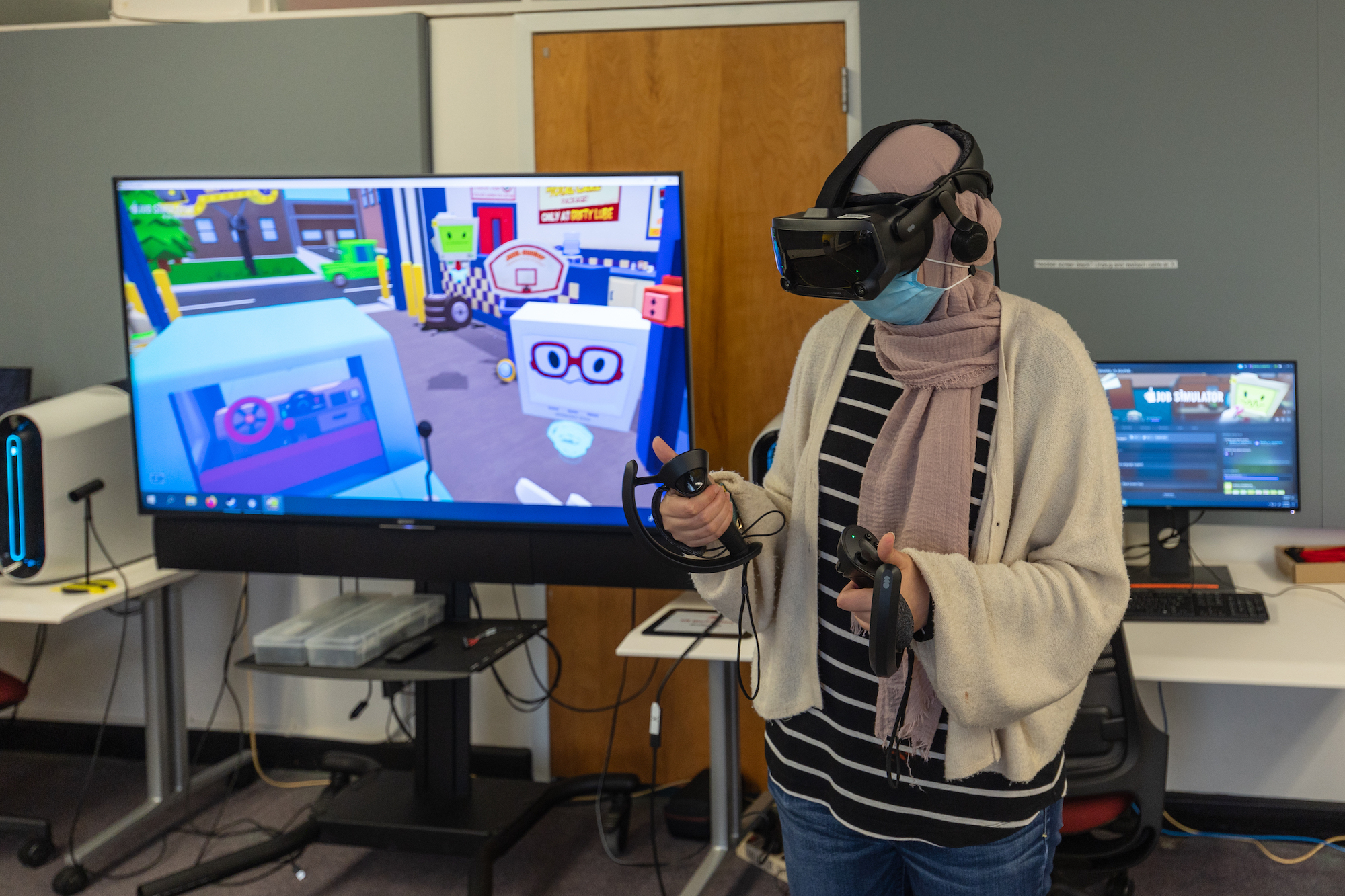 Student wearing a VR headset and holding two controllers, with a screen showing a 3D space behind her with cartoon characters in it